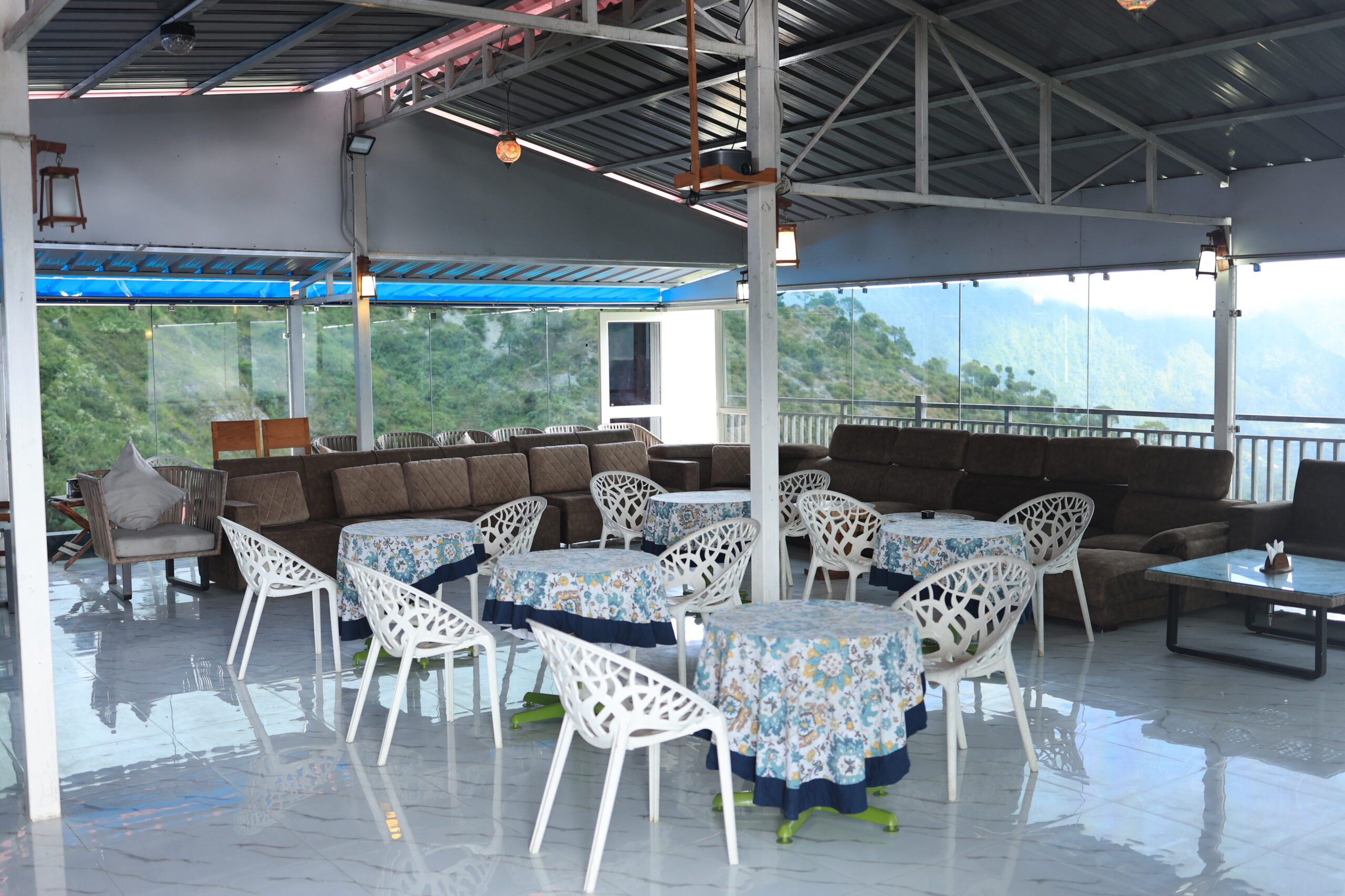 Open-air lounge area at Shivalaya Resort with white chairs, floral tablecloths and mountain view through glass railings.
